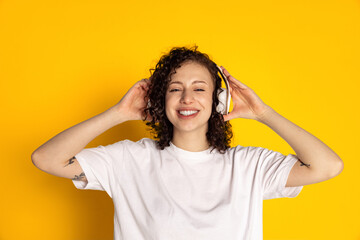 Front view of smiling young beautiful girl in headphones listening to music, audio books isolated on yellow background