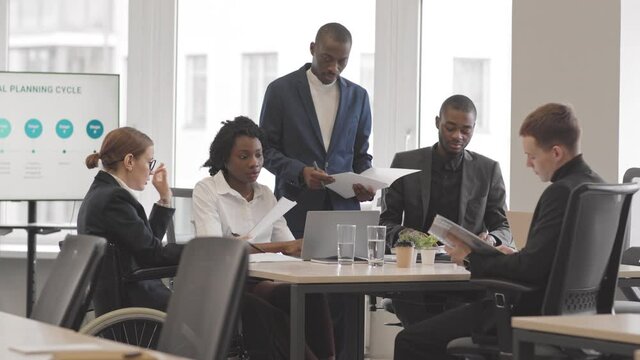 Medium long of female manager in wheelchair, two African American men and woman, and young male Caucasian coworker working on business project in conference room at daytime