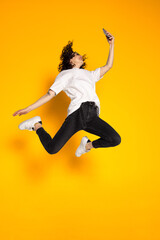 Fototapeta na wymiar Full-length portrait of young cheerful girl, student in white t-shirt and jeans jumping isolated on yellow background.