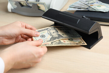 Woman checking dollar banknotes with currency detector at wooden table, closeup. Money examination...