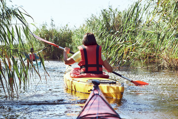 Rear view of a people rowing in a kayaks against a background of green reeds (bulrush) in small river. Summer kayaking in wilderness.