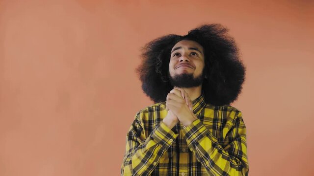 A young man with an African hairstyle on an orange background crosses his fingers and prays. Emotions on a colored background.