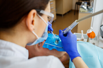 Male patient sits on the dentist's chair and receives teeth restoration, female dentist at the dental clinic. Selective focus