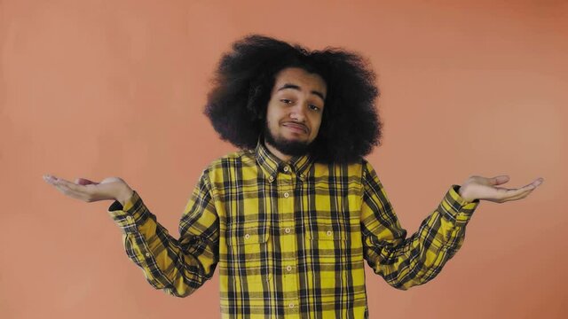 A young man with an African hairstyle on an orange background shrugs his shoulders. Emotions on a colored background.
