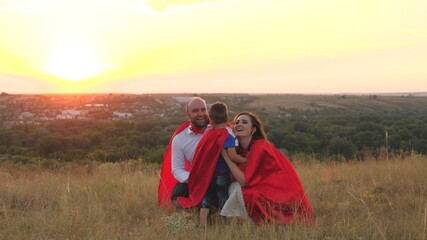 Fototapeta na wymiar mother and father kiss and hug little child sunset, playing superheroes dressed red cloaks, happy family, brave savior costume, fantastic childhood dream heroes, fearless kid boy winner fun play game
