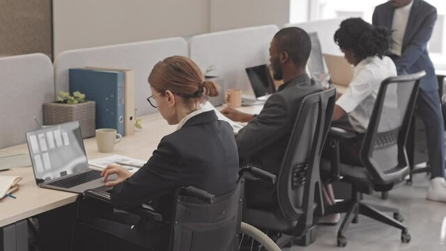 Rear view of short-haired Caucasian woman in wheelchair wearing formal suit, using portable computer, sitting at desk in office next to African American colleagues