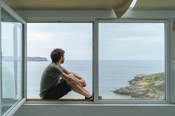 Panoramic rear view of unrecognizable man sit on a window frame looking at the sea. Horizontal view of man isolated at home with a view from the window of the blue ocean on the background.