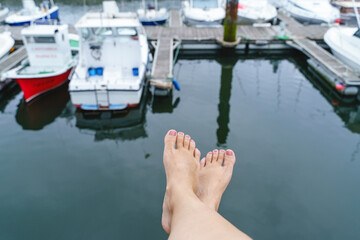 Obraz premium Top view of unrecognizable woman bare feet in Suances harbor. Horizontal view of woman feet isolated with white boat and blue sea background. People and maritime travel concept.
