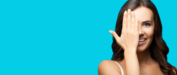 Portrait studio photo of happy smiling brunette woman with one eye, closed by hand, covering part of her face, isolated over aqua blue color background. Copy space area.