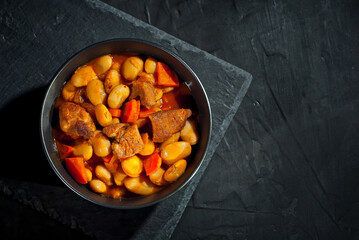 Yellow beans in a black bowl. Boiled beans with meat on a stone table. Beans with tomato sauce and pork. Contrasting dramatic light as an artistic effect.