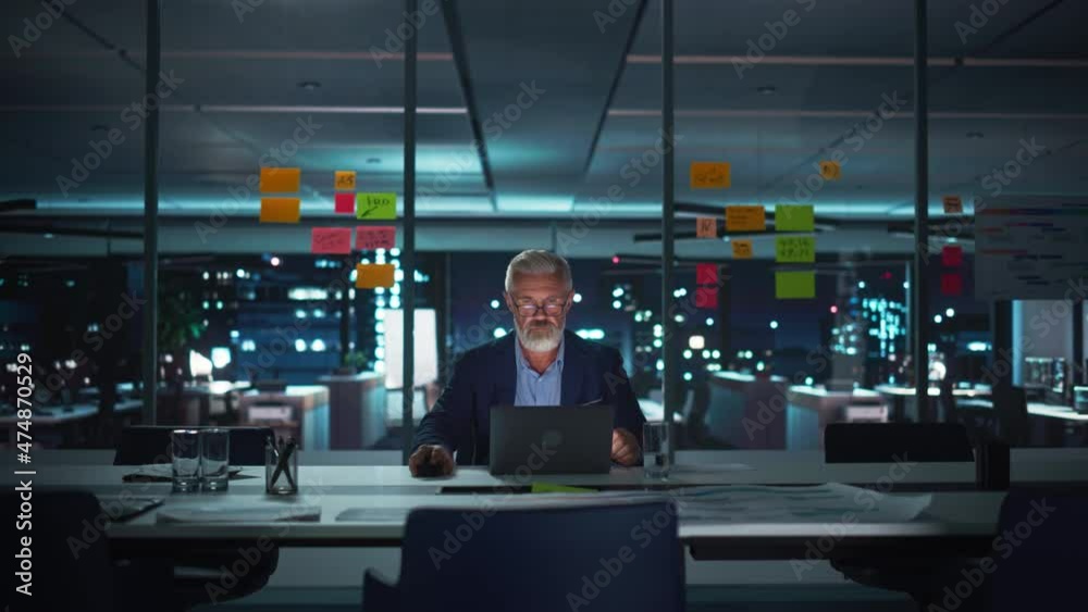 Poster successful senior businessman working on laptop computer in big city office late in the evening. inv