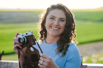 Close Up portrait of smiling beautiful girl taking photo on retro camera. A bright girl in a blue dress is smiling and holding a retro camera in her hands. Girl with a camera at the cottage