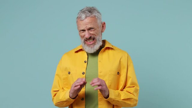 Scared shocked elderly gray-haired bearded man 50s wears yellow shirt look camera covering hiding face with hands peep through fingers isolated on plain pastel light blue background studio portrait