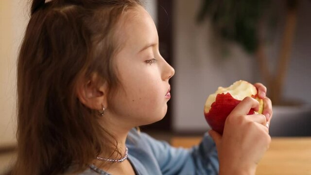 Close up portrait of happy little girl biting ripe delicious red apple, at kitchen interior, slow motion, healthy eating concept. Child with snack fruit
