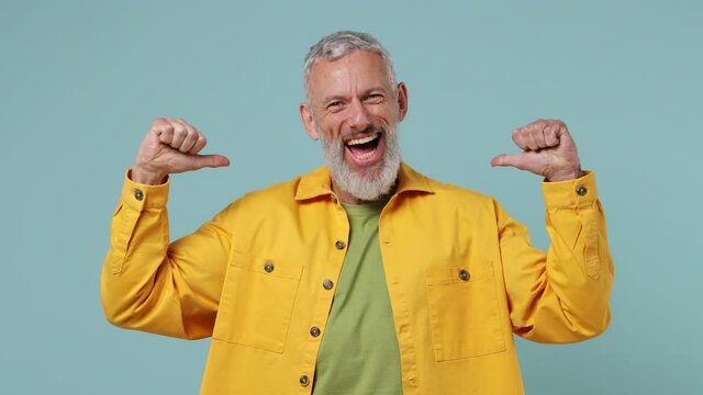 Confident fun elderly gray-haired bearded man 50s wears yellow shirt pointing fingers on himself blinking showing thumb up like gesture isolated on plain pastel light blue background studio portrait