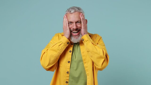 Happy fun elderly gray-haired bearded man 50s wears yellow shirt look camera surprised ask what wow omg no way say yes put hands on face isolated on plain pastel light blue background studio portrait