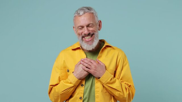 Smiling good kind surprised excited elderly gray-haired bearded man 50s wears yellow shirt ask who me oh it so sweet put hands on chest isolated on plain pastel light blue background studio portrait