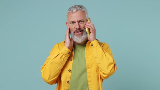 Happy calm fun elderly gray-haired bearded man 50s wears yellow shirt hold use talk on mobile cell phone conducting pleasant conversation isolated on plain pastel light blue background studio portrait