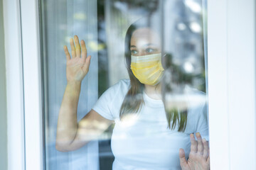 Young woman in quarantine wearing a protective mask and looking through the window