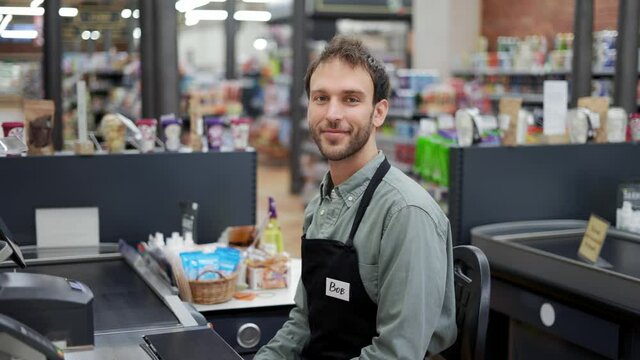 Handsome man in supermarket waiting at cash desk for next customer. Smiling cashier in black apron