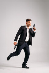 A young guy in a black suit with a gun in his hands on a white background in the studio, he depicts a security guard, bodyguard or Agent