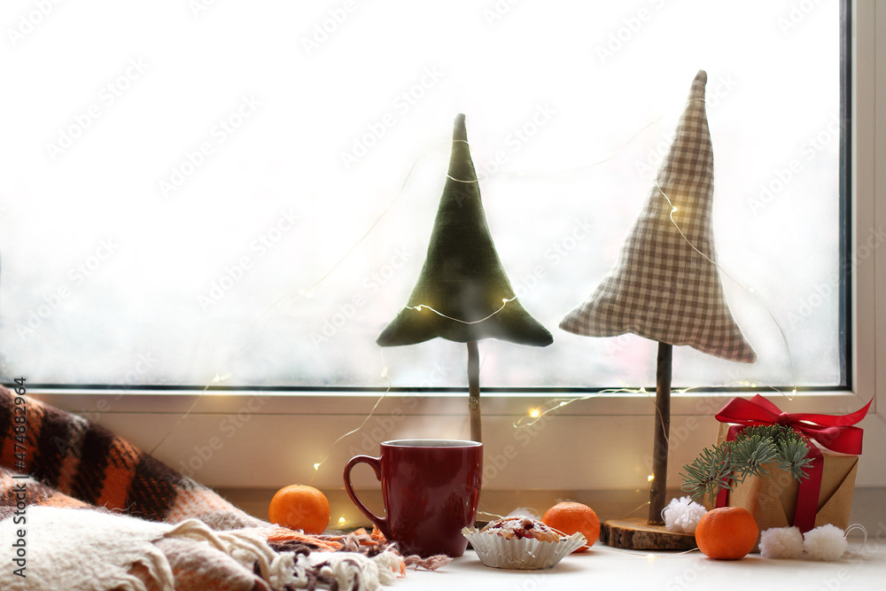 Wall mural hot drink in a mug and berry cupcake on the windowsill with a plaid, tangerines, a gift, decorative Christmas trees on the background of the window. cozy winter Christmas morning