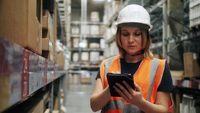Female worker warehouse wearing hard hat and safety vest checks stock and inventory with tablet using e-commerce app, standing near shelves with cardboard boxes. Logistics concept.