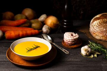 Carrot and potato soup with rosemary twig on a grey wood table with a spoon, onion, potatoes, carrot, garlic, bread loaf, pork belly, laurel and rosemary.