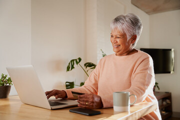 Elderly multi-ethnic female smiling while on an online call. Sitting with laptop at kitchen counter. 