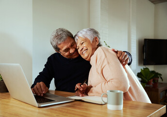 Multi-cultural elderly couple celebrating after receiving good news. Sitting at modern kitchen counter with laptop.