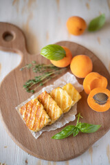 Slices of fried halloumi cheese with a beautiful grilling pattern rests on a wooden tray with apricot slices.