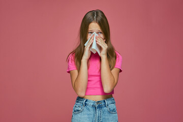 Sick girl child cry, blowing runny nose, sneezing in tissue, suffer seasonal allergy or flu symptoms on pink background