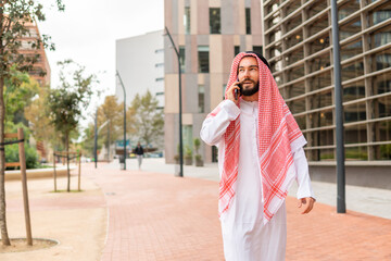 Arab businessman in traditional clothes making call on mobile phone while walking city street