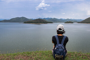 Back view of a young asia woman sitting and enjoying peaceful moment of beautiful view at lake shore with mountains range in background. Holiday travel concept, Rest and enjoyment, lifestyle, 