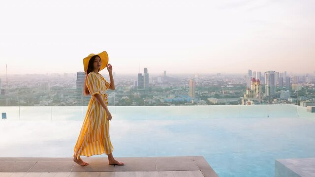 Slow motion - Beautiful woman walking on the edge of the pool wearing a long yellow dress and yellow hat. Female walking by the infinity swimming pool and background city scenery with sunset.