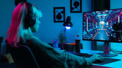 Isolated Beautiful Caucasian Blonde Gamer Girl Playing Video Games on Her PC Computer, Looking Focused and Serious as She Plays Around. Wearing Black Headphones, Sitting in Her Dark Moody Bedroom. © Stock Unit