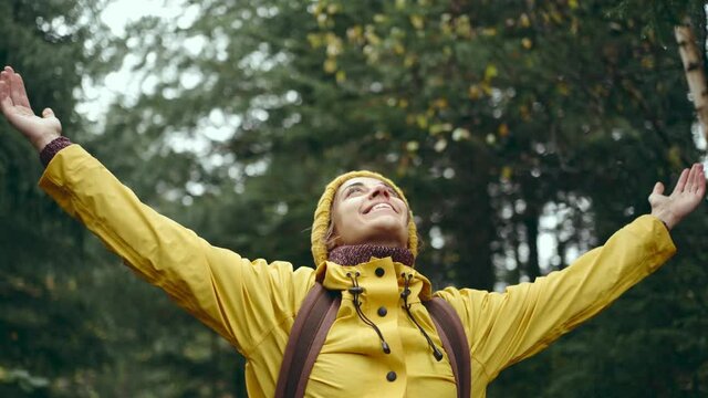 portrait of inspired young pretty woman hiker with backpack, wearing bright yellow raincoat and beanie looks up raising hands enjoying nature beauty, freshness and freedom in wonderful autumn woods.
