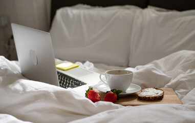 Morning coffee in bed. Cup of delicious, aromatic coffee, sandwich and two ripe strawberries on wooden kitchen board lying near laptop on white bed. Beakfast at home-office in time quarantine.
