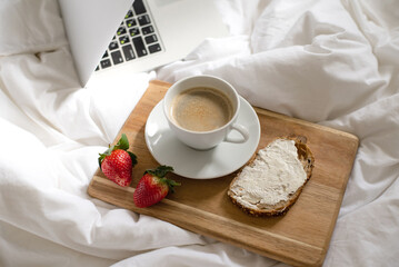 Morning coffee in bed. Cup of delicious, aromatic coffee, sandwich and two ripe strawberries on wooden kitchen board lies near laptop on white bed. Beakfast at home-office in time quarantine.