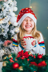 Little smiling girl Santa helper holding christmas mug with hot chocolate and marshmallows with christmas wreath on background