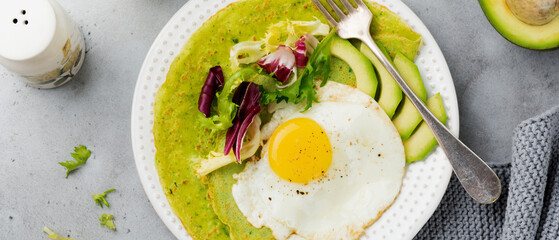 Spinach green crepes (pancakes) with fried egg, avocado and leaves of mix of salad on ceramic plate on gray concrete background. Concept of healthy breakfast. Selective focus. Top view. Copy space.