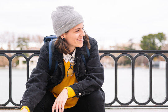 Smiling woman in casual clothes and backpack sitting on embankment