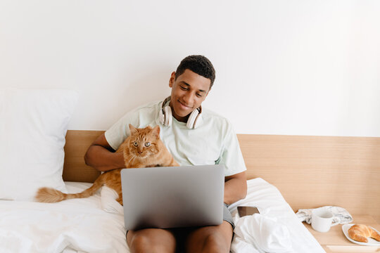 Black man wearing t-shirt petting his cat while working with laptop