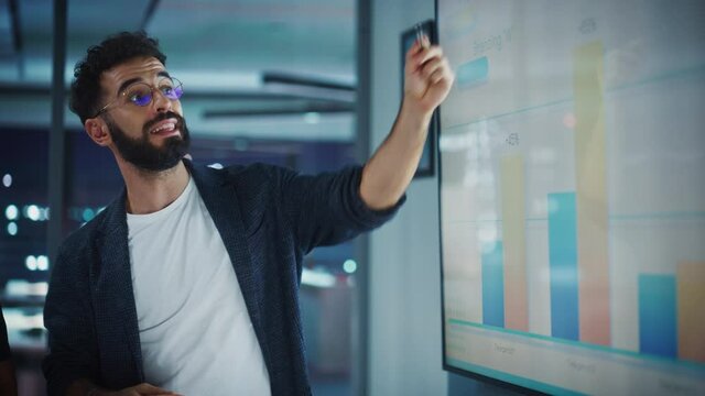 Company Operations Director Holds Sales Meeting Presentation for Employees and Executives. Creative Male Uses TV Screen with Growth Analysis, Charts, Ad Revenue and Data. Work in Business Office.