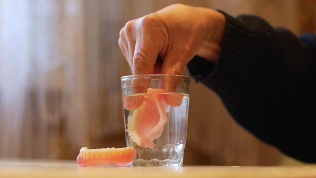 The topic is pensioner and false dentures. On the table is a glass of water in which an elderly man puts his removed dentures. The problem of tooth loss. Installation of removable dentures.