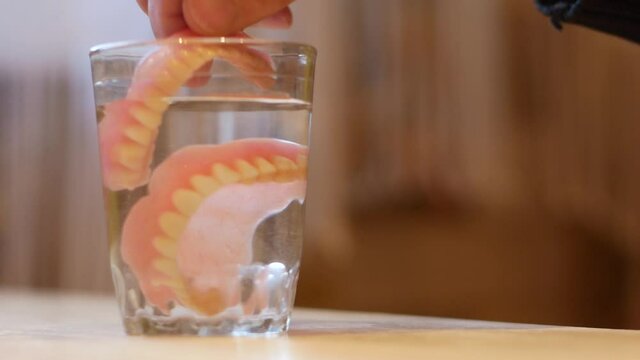 Close-up of a glass of water into which false teeth are immersed in dentures. An elderly person who has lost his teeth. Dental implantation clinic. Topic of tooth loss and prosthetics. selective focus