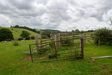 broken wooden fencing and metal gate with Devon hills and grazing land in the background on a cloudy day
