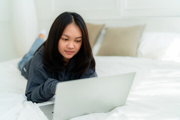 Fototapeta na wymiar Portrait, a cute Asian teenage girl. wearing gray shirt lying in bed with a notebook computer in the bedroom to study online with a happy face
