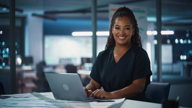 Portrait of a Successful African American Businesswoman Working on Laptop Computer in Big City Office Late in the Evening. Happy Black Female with Curly Hair in Stylish Dress Smiles on Camera.