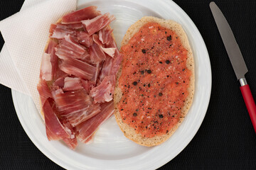 Plato blanco, con tostadas de jamón ibérico y tomate triturado, sobre fondo negro, vista superior